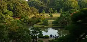 A pond in a wooded park with a bridge and grassland.