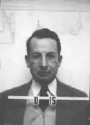 Mug shot of a man with glasses in suit and tie. The number O-15 appears in front of him.