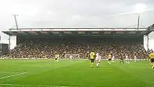An impressive grandstand behind a goal net, filled with people, viewed from the other end of the stadium.