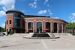 On the left is a two-storey red brick rotunda with a full-height five-pane wide dark window showing parts of the interior atrium, and at which base is the main entrance to the theatre. To the right is a red brick wall arcing away, with nine identical and equally spaced four-pane wide windows stretching from the ground to nearly the roof. It is fronted for its entire length by a stairway with about seven steps, leading to a piazza in the foreground.
