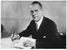 Black and white photograph of a man wearing suit holding a pen with one hand and a piece of paper with the other.