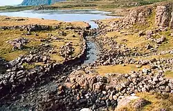 A stone lined ditch of primitive construction leads from a small lake. Rocky heathland lies on either side and there are tall cliffs in the distance.