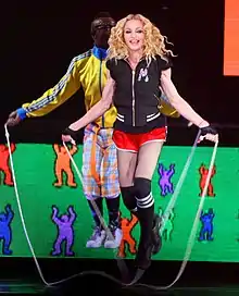 Picture of a blonde woman jumping rope. She's wearing a black vest, red shorts, and black knee-high socks. Behind her, a black male wearing a yellow-and-blue sweatshirt, and blue-and-orange pants, also jumps rope. The backdrop behind them is green and depicts different colored human-like drawings.