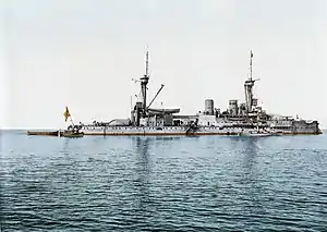 A large gray battleship with two tall masts sits idly in calm waters. Three small boats are tied alongside.