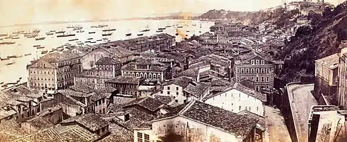 Photograph looking down a hillside overlooking the buildings of a town with a harbor beyond which is crowded with various vessels