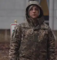Ashton-Cirillo in uniform, her hood pulled up over blonde hair, wearing a solemn expression, in front of a gray building. The camouflage uses a pixelated style. The name on the right side of her chest reads "BLONDE". The Ukrainian flag and the patch of the Noman Çelebicihan Battalion are visible on her right shoulder.