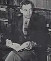 A photograph of Saul Bellow with an open book before a bookcase. He is wearing a suit and has somewhat curly hair.