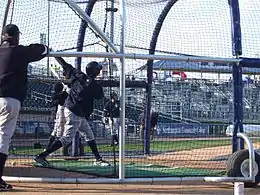 A man in gray baseball pants and a navy blue top swinging a bat at a baseball in a batting cage
