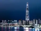A nighttime shot of the Lotte tower from across the river. The tower is lit up and is far taller than any of the apartment buildings around it. By the riverside, a row of streetlights are shining on the river.