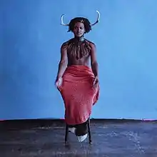A photo of the artist sat on a stool in the middle of a room with blue walls and a dingy concrete floor. Shamir has animal horns coming out of his hair and furry, hooved feet, and is wearing an ankle-length pink skirt, no shirt, and traditional African neckwear with a small red heart adorning it around the top of his left pectoral.