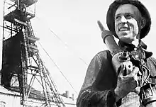 Alexei Stakhanov and another man at work in a Soviet coal mine. Stakhanov, while holding a drill, is seated at the coal face, his head turned to speak to his colleague.