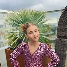 Mansfield smiling for the camera in a glittering purple-pinkish dress before attending the Children's & Family Emmy® Awards as a presenter in 2022. She is on a rooftop with the city of the Hollywood in the background.