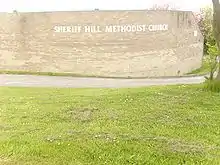Behind a large area of mown grass stands a beige brick wall which curves around a corner to the right. The wall bears the words "Sheriff Hill Methodist Church", which follow the curve of the wall.