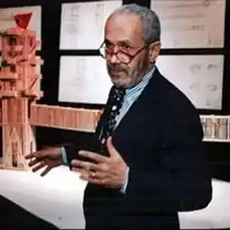 Waist high portrait of man with greying hair wearing dark suit, light blue shirt and black rimmed glasses, speaking in front of apparently wooden sculpture with long bridge appendage