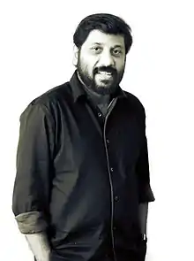 Upper-body photo of smiling man with dark hair, mustache and beard wearing dark-colored button down shirt