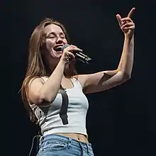 A woman wearing white tank top and jeans, holding a microphone, with one hand up to her chest while smiling.
