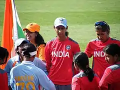 Eight female cricketers stand on a field. Two players are in red practice jerseys; three other players in red jerseys are facing toward them; a player in a blue shirt is facing away and hides another player in a blue game shirt. In the upper left corner is a fan with an Indian flag