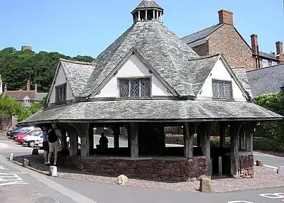 A small single-story building with a pyramid shaped roof, to the side of a road lined with buildings. Some private small cars visible. Trees in the distance with the skyline of Dunster Castle.
