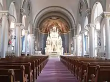 Looking down central aisle with pews on either side; line of pillars connected at tops by arches on either side; altar under half-dome at far end