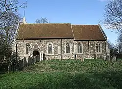 A plain stone church with a red tiled roof seen from the south, with the top of a small spire seen protruding above the left of the nave