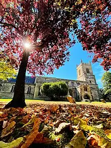 A photo of Bicester's St Edberg's Church, a Cotswold stone building, in the sunshine, with the sun shining through a tree.