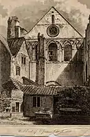 Monochrome illustration of the west end of St James' church, published in 1820. The image shows the parts of the priory stonemasonry incorporated into the church facade and a jumble of non-ecclesiastical buildings in the foreground including a tall chimney.