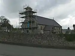 A small stone church with a slate roof see from the southwest. Scaffolding surrounds the west end and bellcote, and the porch