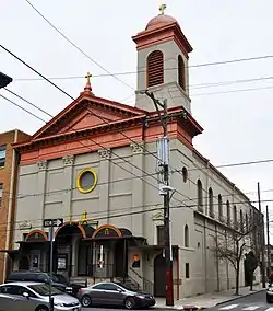 A tan church with red and gold accents stands among parked cars and overhead wires on an urban street corner