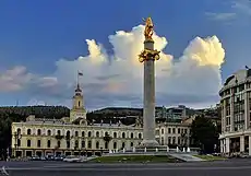A city square; at the center is a large pillar with a golden statue on top; in the background is a large building with a steeple.