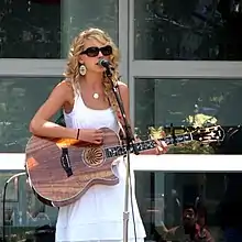 Taylor Swift, donning a white sundress and large sunglasses, playing a wooden acoustic guitar