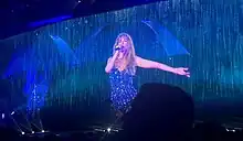 Swift, dressed in a sparkling purple dress, standing onstage as a male dancer holds an umbrella besides her