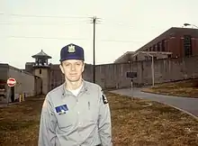 Ted Conover in his late 30s, standing outside of the prison perimeter wall wearing his correctional officer uniform hat and button-up shirt, with officer’s notebook and pen in his shirt pockets