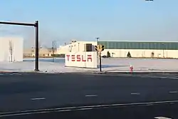 Large white sign, with red letters spelling "TESLA", in the parking lot of a long, low building.