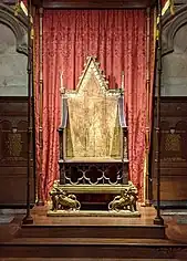 An old gilded wooden chair on a raised platform in front of a red curtain.