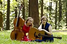 Two women sitting in woodland with a cello and a guitar