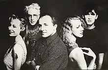 Five adults framed in upper body. In the front row, the left female is shown in left profile, slightly turned to her left and smiling, she wears a white dress with black polka dots and has a serpent tattoo on her upper left arm. Behind her, a slightly balding male is more turned towards the front and has his arms folded across his chest, his shirt is dark with white polka dots. Back to back to him is the second female in right profile with her right arm touching her shoulder. In the back row, the left male has white hair and is facing forward, he is wearing glasses and has an obscured design on his shirt. The right male has dark hair, he is staring forward and wears a black tee shirt.