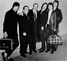 The Kinks and the two executive, all in suits, pose for a group photo, luggage in hand.