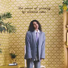 Alessia Cara, wearing a grey suit, stands afront a cream-coloured background with the album title and her name displayed above her