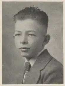 A white teenaged boy with dark hair; he is wearing a suit coat and tie, and is facing the camera's left.