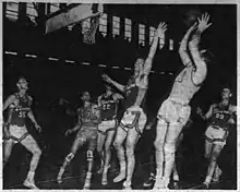 Newspaper photograph of an American Basketball League game between the Scranton Miners and Elmira Colonels. Leroy Chollet is jumping with one hand raised to contest a jump shot from Danny Finn who is fading away with the ball raised above his head. Three other players position themselves for a rebound near the basket.