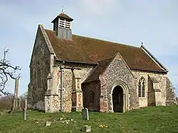 A small stone church with a red tiled roof seen from the southwest, showing a south porch and a small bellcote