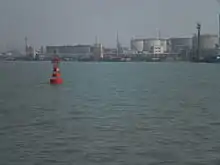 A red port-side buoy with an oil wharf and tankers in the background