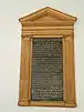 A photograph of a memorial in a wooden frame hanging on the wall of a church. The frame is a carved depiction of a Greek temple with columns and a small gable. The inscription is of gold lettering on a black background