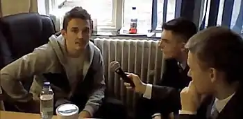 Two schoolchildren can be seen at the right, one of them holding a microphone to a man peering at the left.