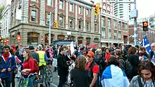 A photograph of a group of people protesting in front of a large, brown-bricked building with grey columns and many grey-rimmed windows