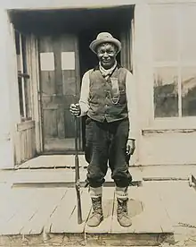 Tonené standing on a porch, holding a rifle in his right hand.