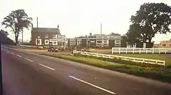 A faded period photograph of the motel on a grey day.  The main road is in the foeground, from lower right to half way up the left side.  In the middle distance a two-story 18th or 19th century building stands at right angles to the road, it's windowed frontage facing our vantage point.  It is on the opposite side of the road to us, beyond the entrance track.   This side of the track, in the centre of the photograph and still on the opposite side of the road to us are a range of low, flat-roofed, single story buildings, almost certainly new at the time.  There is low whir post-and-rail fencing on the verge this side of the motel.  The older building is probably red brick. The new buildings have dark grey walls.  The sign reading 'Trenton Motel' is clearly visible by one of the modern buildings.