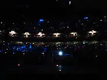 A darkened concert venue. People are in multiple levels of seating, and tiny lights are scattered throughout the audience. On the left, performers stand on a stage lit with blue lighting.