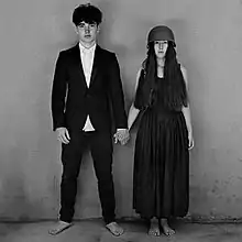 A monochrome image of barefoot young man in a dark suit coat holding hands with a barefoot young woman in a dark, ankle-length dress who is wearing a military helmet