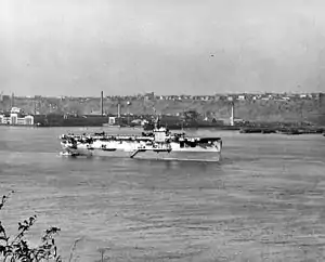 A World War II-era aircraft carrier at anchor. Several aircraft are on her flight deck and land and buildings are visible in the background.